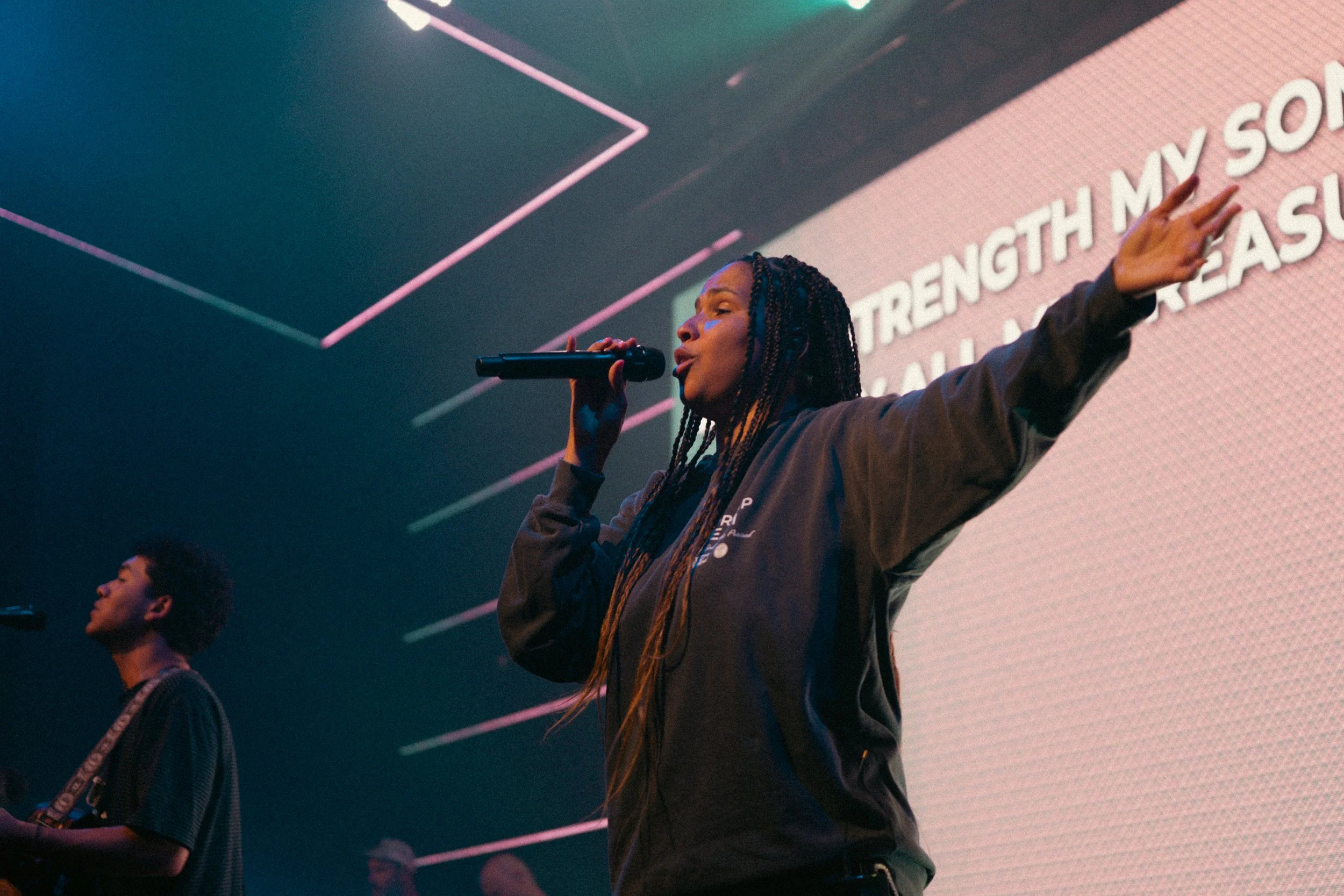 A woman with braided hair is singing into a microphone on stage with her arm raised. Behind her is a large screen with partially visible white text on a pink background. A man and another person are also present on stage.