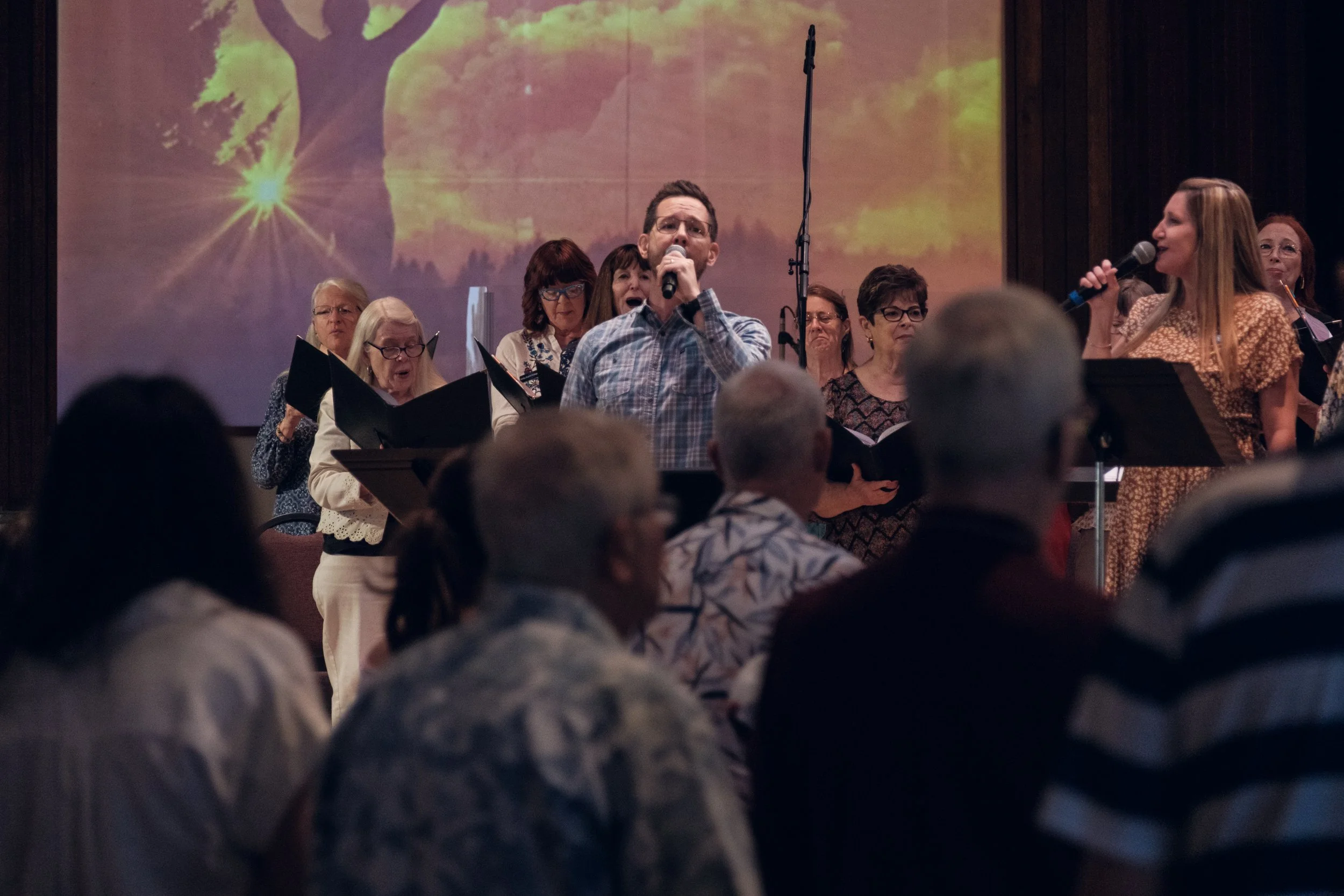 A group of people on stage, some holding choir folders, with a man in plaid shirt speaking into a microphone. A woman on the right is also holding a microphone, with a large screen in the background showing a tree with sunlight.