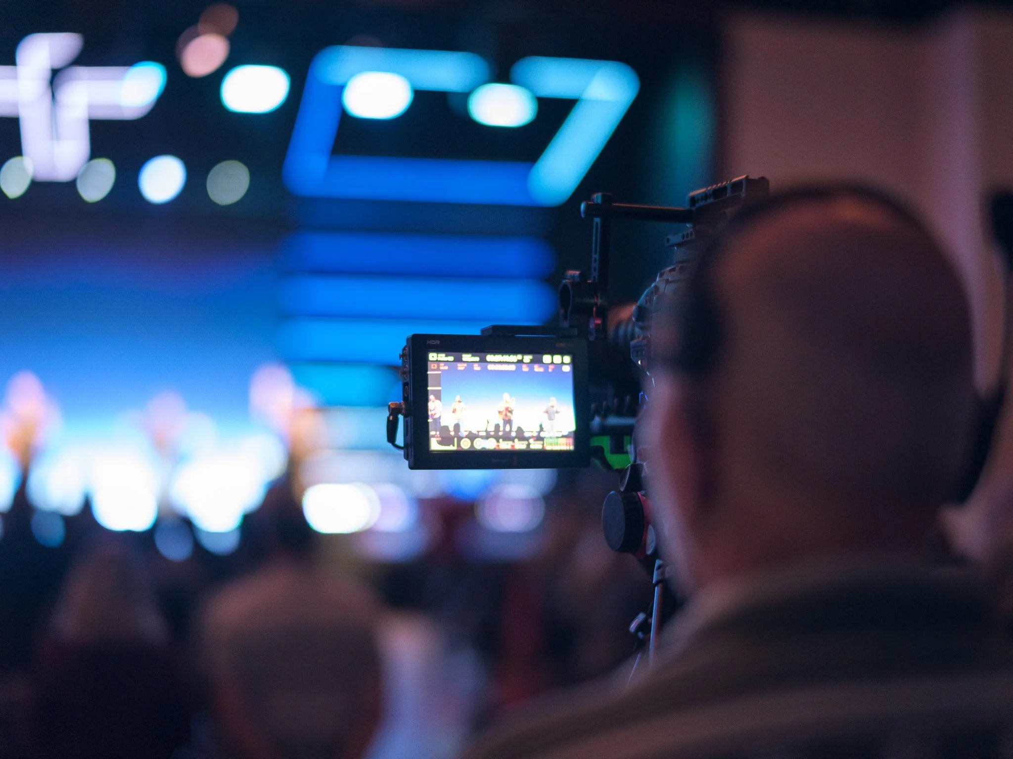 A camera with a display screen capturing an image of a stage with performers, in a dimly lit setting with colorful lights.