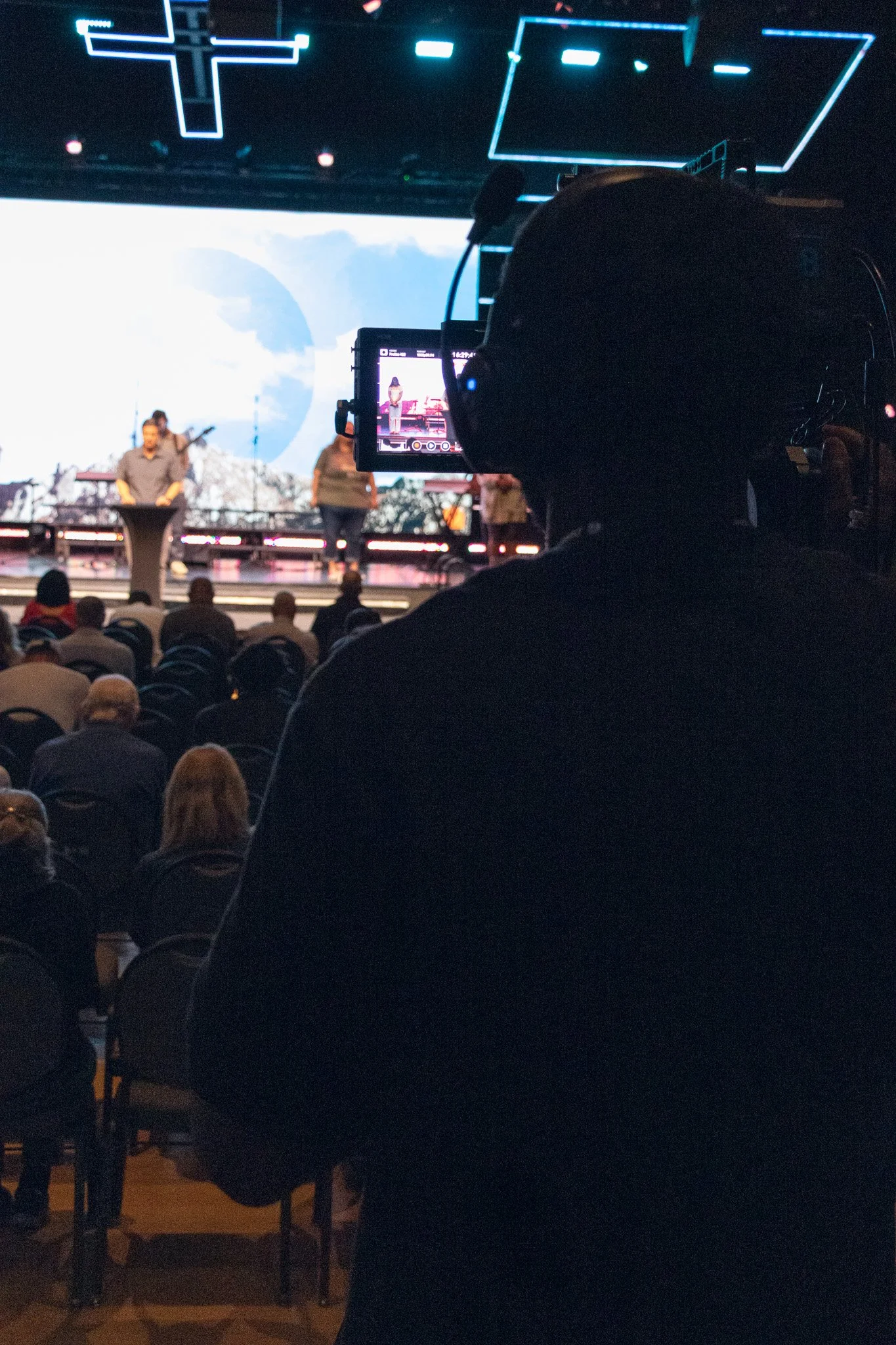 A person in the foreground wearing headphones and taking a photo or recording a video with a camera. In the background, on stage, there are two people, one playing a keyboard and the other standing, with a large screen behind them displaying a sky with clouds.