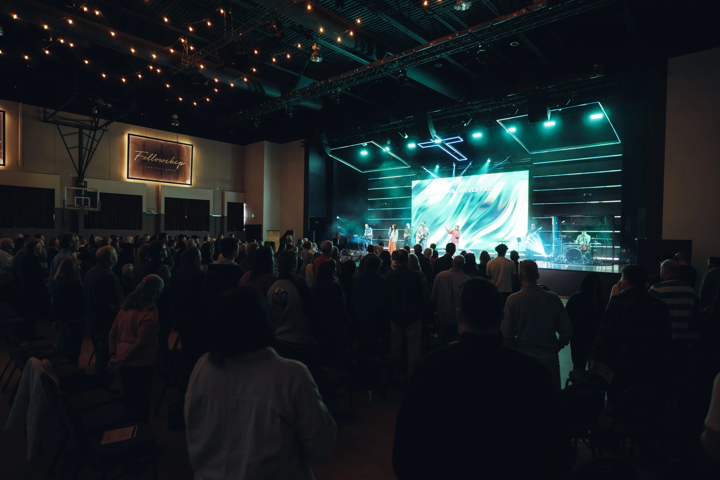 A large crowd of people attending an indoor concert or worship service, seated and standing, facing a stage with a band and a large screen displaying words. The stage is decorated with modern lighting, and hanging string lights add to the ambiance. A sign on the wall reads 'Fellowship Creating Space'.