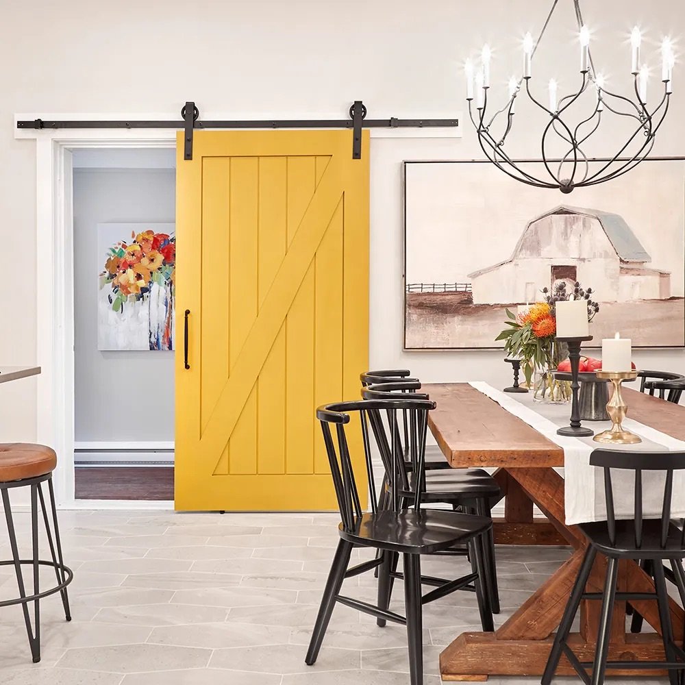 A dining room with a yellow sliding barn door, black chairs around a wooden table, a large chandelier, and artwork on the walls.