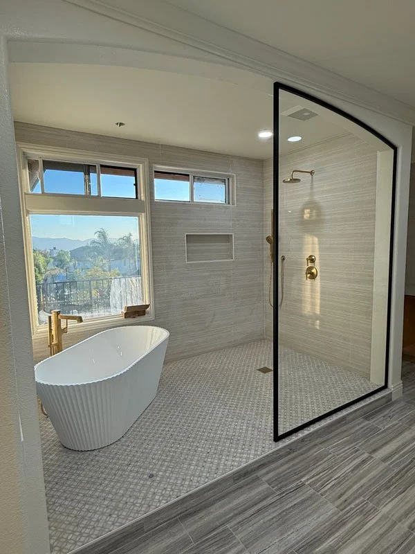 Modern bathroom with a freestanding bathtub, large window, and a glass-enclosed shower with gold fixtures.