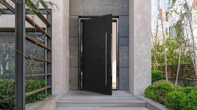 Modern black front door slightly open with silver handle, surrounded by concrete wall, leading to an outdoor area with plants and trees.