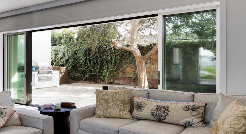 Living room with beige sofa and pillows, large sliding glass doors open to a patio with a barbecue grill, tree, and greenery outside.