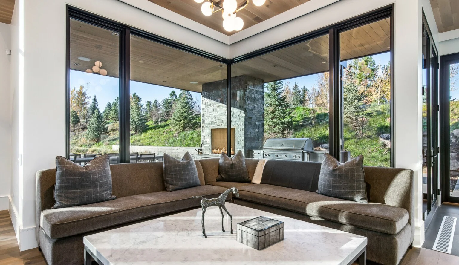 Living room with a large L-shaped sofa, throw pillows, a marble coffee table, and large glass windows showing a patio with an outdoor grill and a wooded landscape.