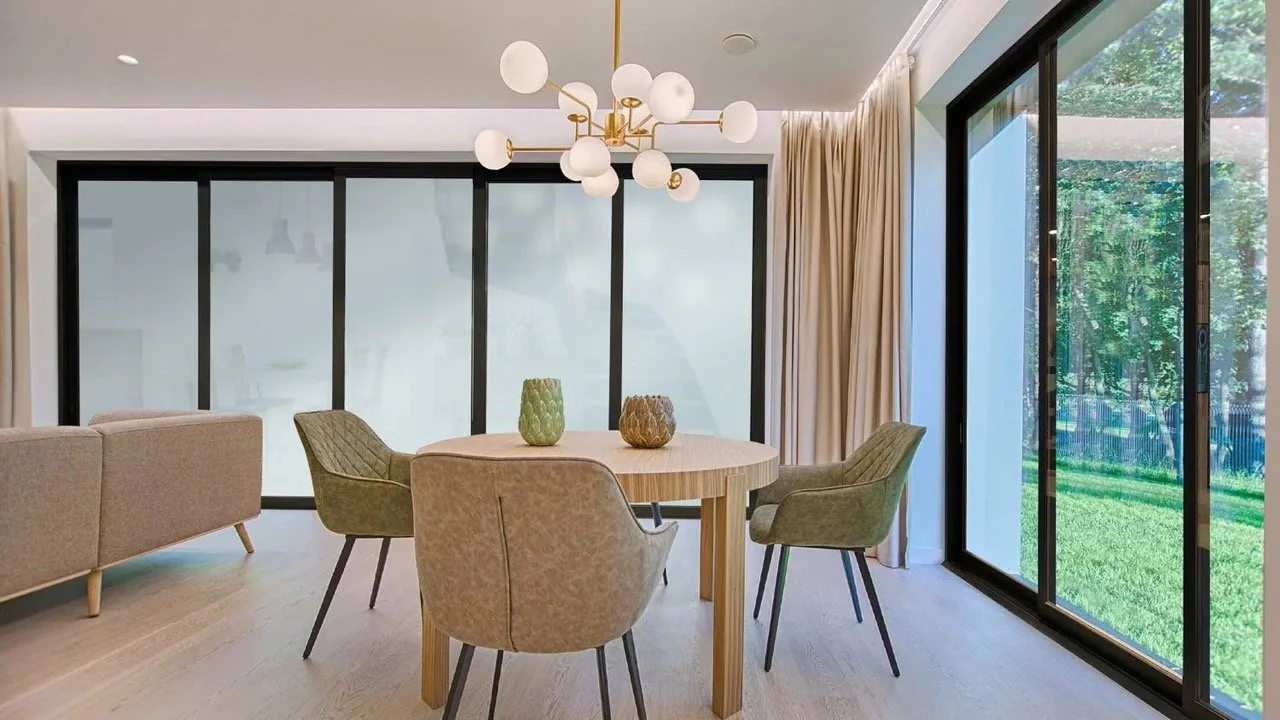 Modern dining room with a round wooden table, four upholstered chairs, large sliding glass doors, a contemporary chandelier, and a view of a green backyard.