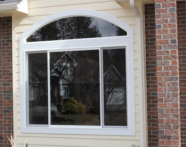 A large house window with an arched top section and three sliding panels, set in a house with beige siding and a brick corner.