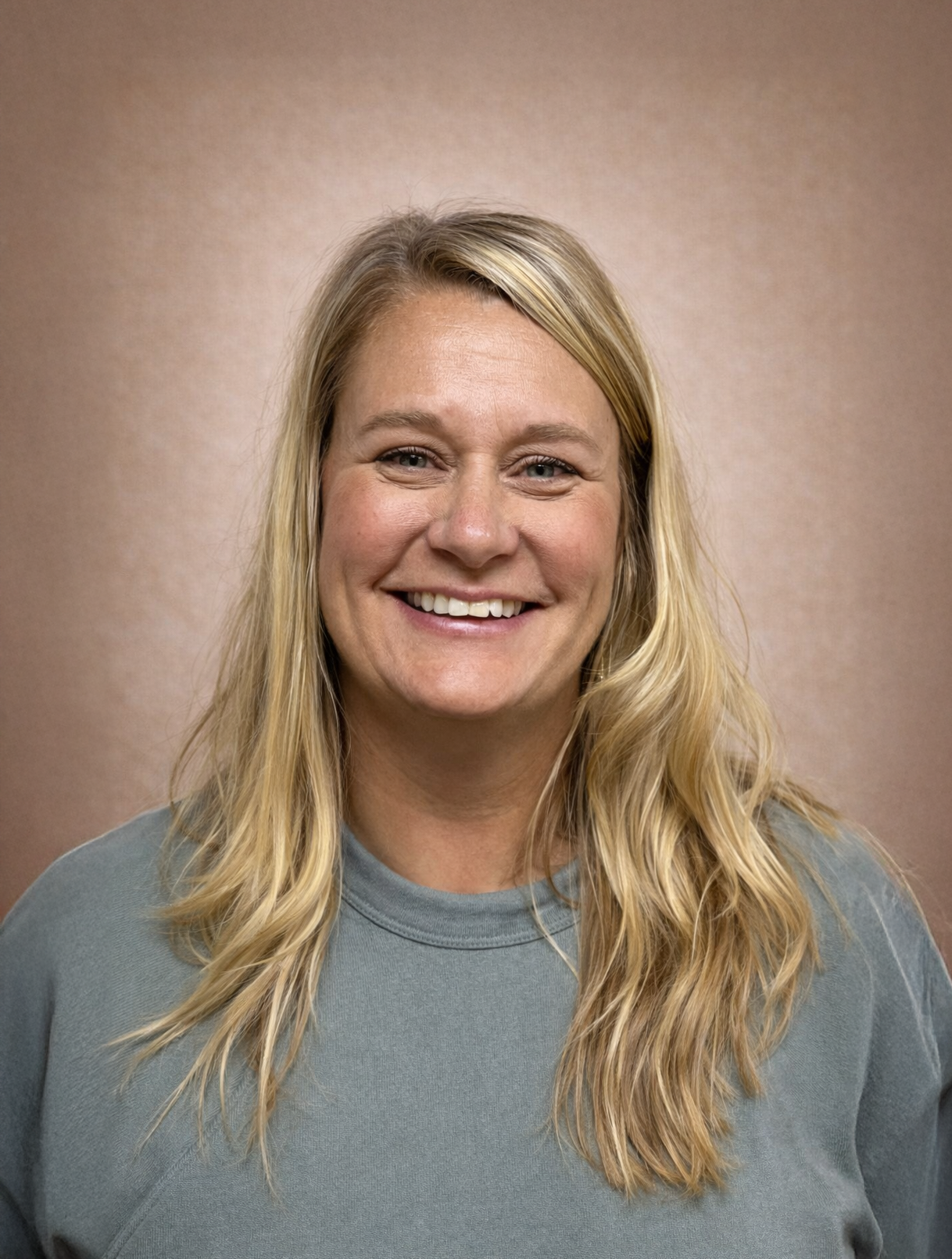A smiling blonde woman with wavy hair wearing a grey top against a brown background.