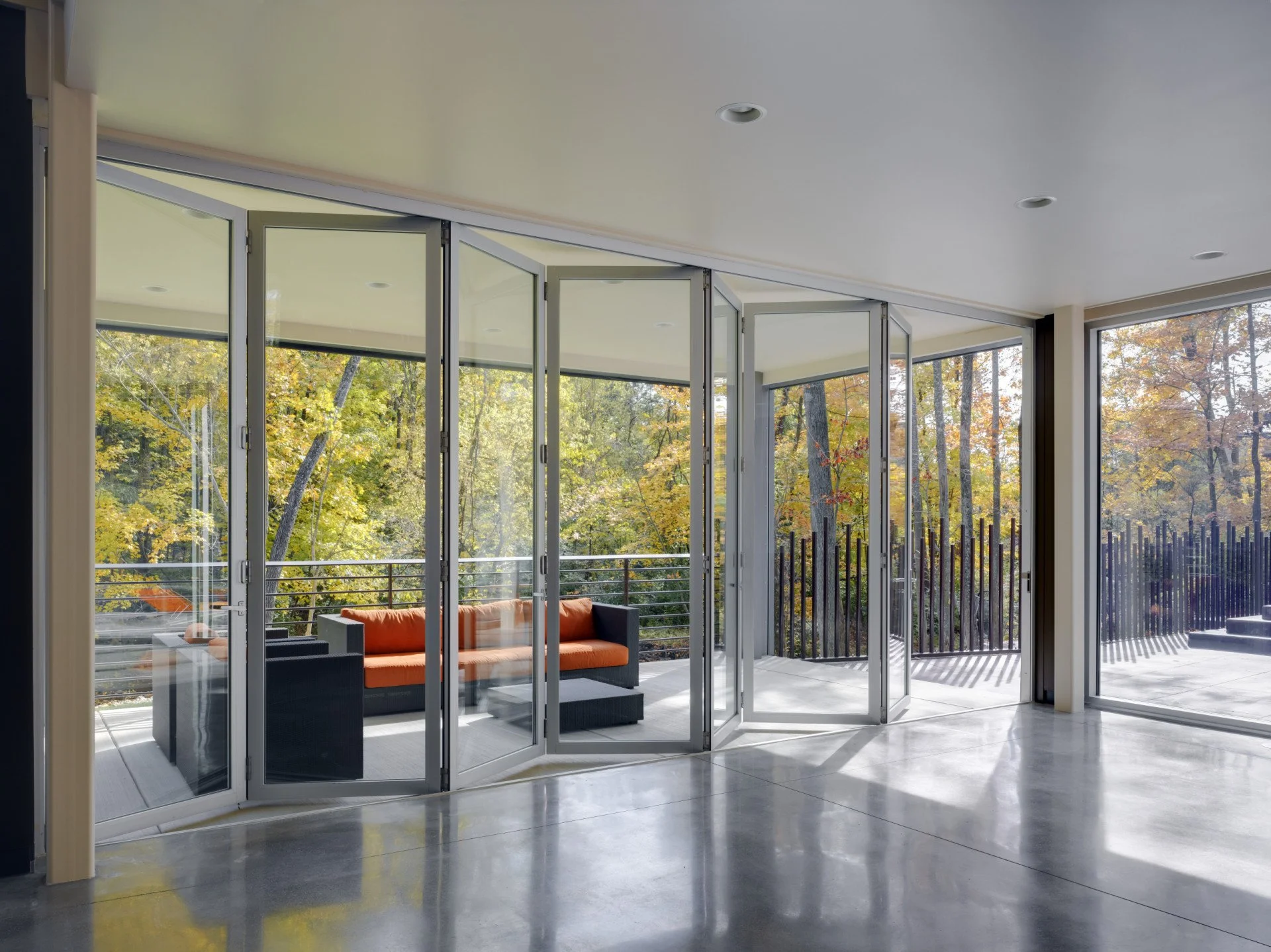 Modern living room with large glass sliding doors opening to a balcony, showing outdoor trees with autumn foliage, and orange couches visible through the doors.
