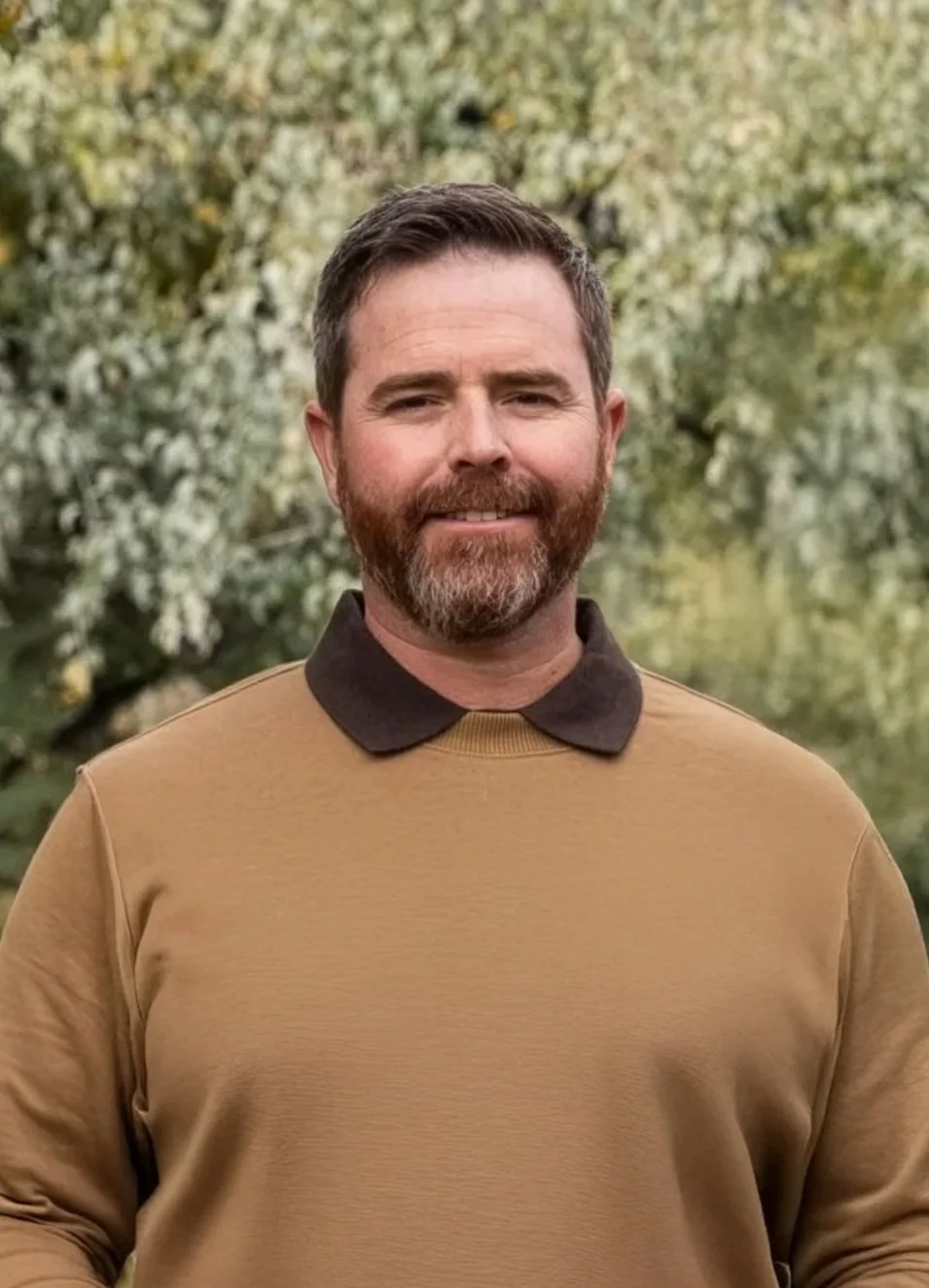 A man with short dark hair and a beard, wearing a tan shirt with a dark collar, standing outdoors with green trees in the background.