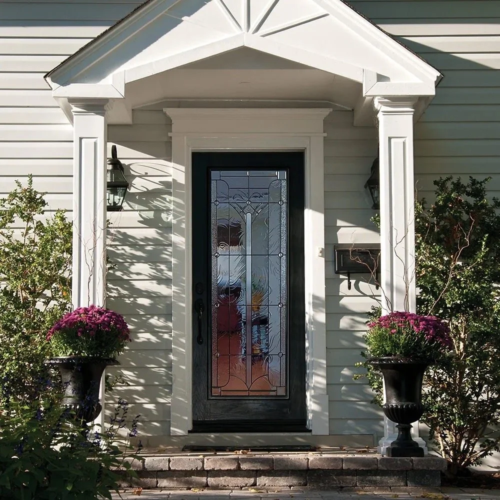 Front door of a house with white siding, decorative glass door, black mailbox, two black outdoor wall lanterns, and two large planters with pink flowers on either side of the entrance.
