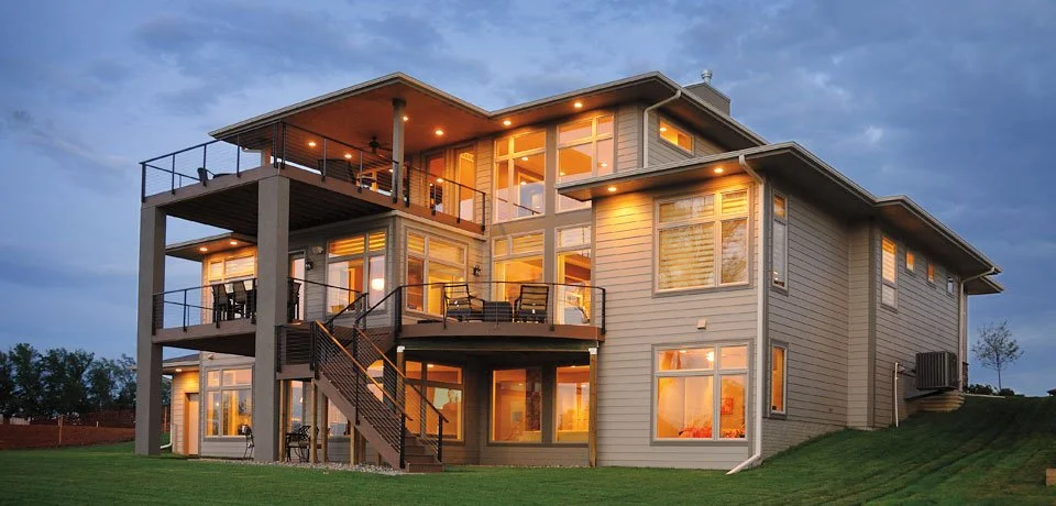 Modern multi-story house with large windows and multiple balconies, illuminated at dusk, on a grassy lawn.