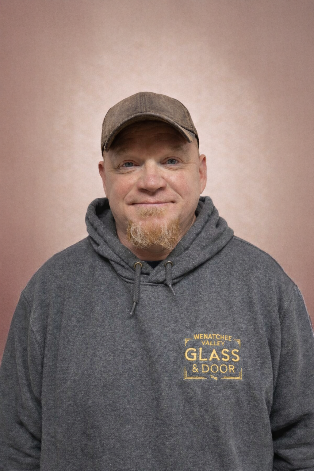 A man with a light complexion, wearing a brown cap and a gray hooded sweatshirt with yellow logo that reads 'Wenatchee Valley Glass & Door,' standing against a plain pinkish wall.