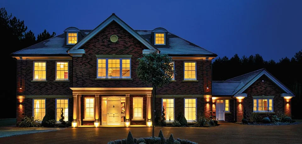 Exterior of a large brick house illuminated at night with lit windows and landscaping.