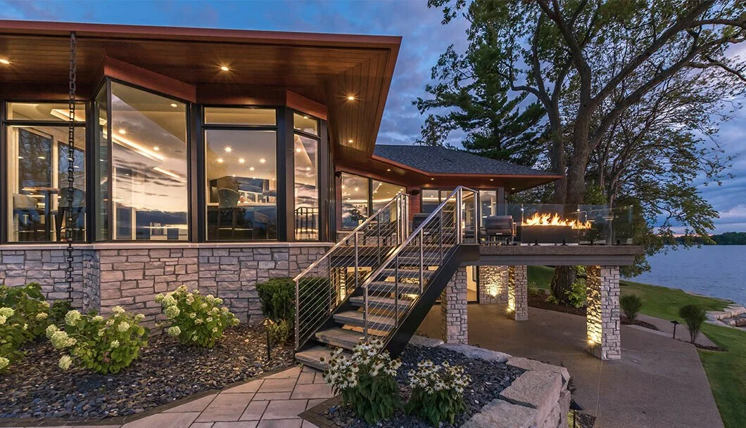 A modern house with large windows and a stone exterior, featuring a lit outdoor fireplace on a deck overlooking the water, with a staircase leading down to landscaped grounds.