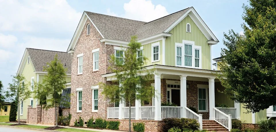 A two-story house with brick and pale green siding, white porch, and trees in front.