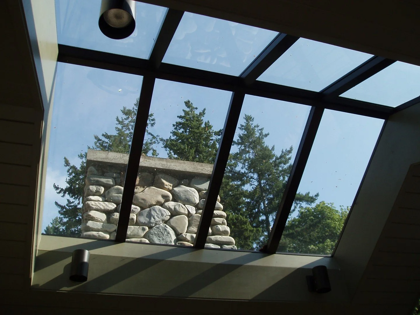 View of a glass skylight window with a stone chimney and green trees outside.
