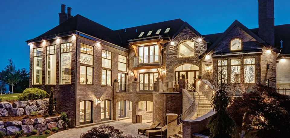 Large stylish brick house illuminated at night with multiple windows, balconies, and outdoor stairs, surrounded by landscaped yard.