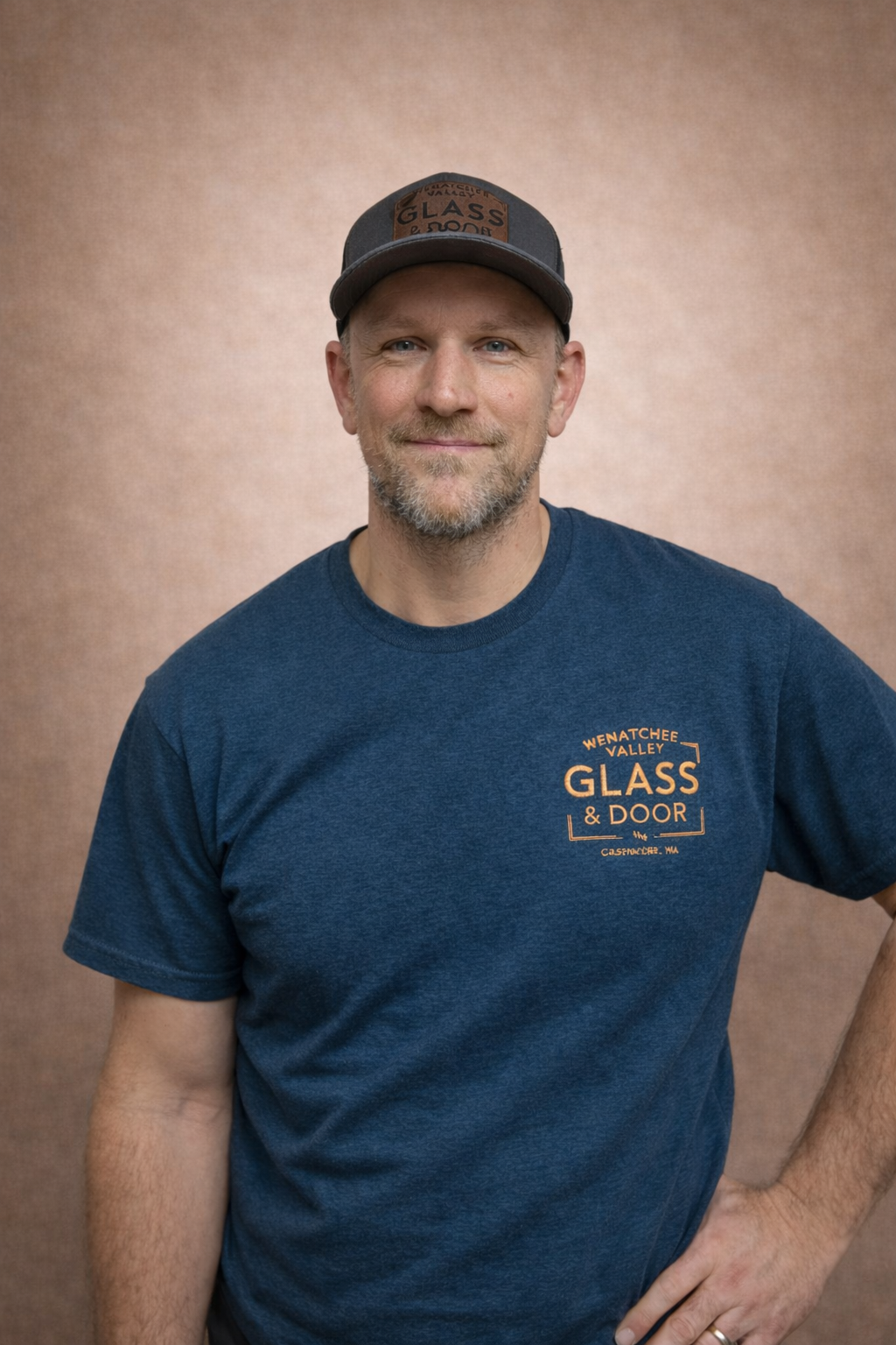 A man wearing a dark blue T-shirt with orange text that reads "Wenatchee Valley Glass & Door" and a baseball cap. He is standing against a brown background, smiling.