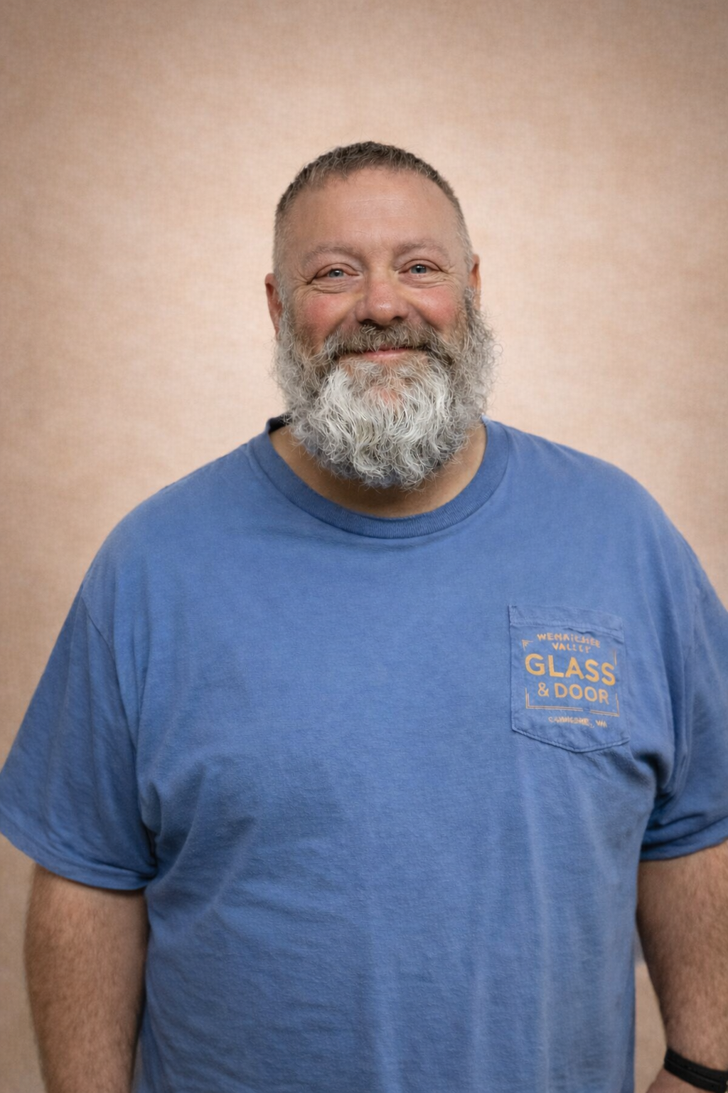 A middle-aged man with a full beard and short hair, wearing a blue T-shirt with a pocket that reads "Wenatchee Valley Glass & Door," standing against a beige background.