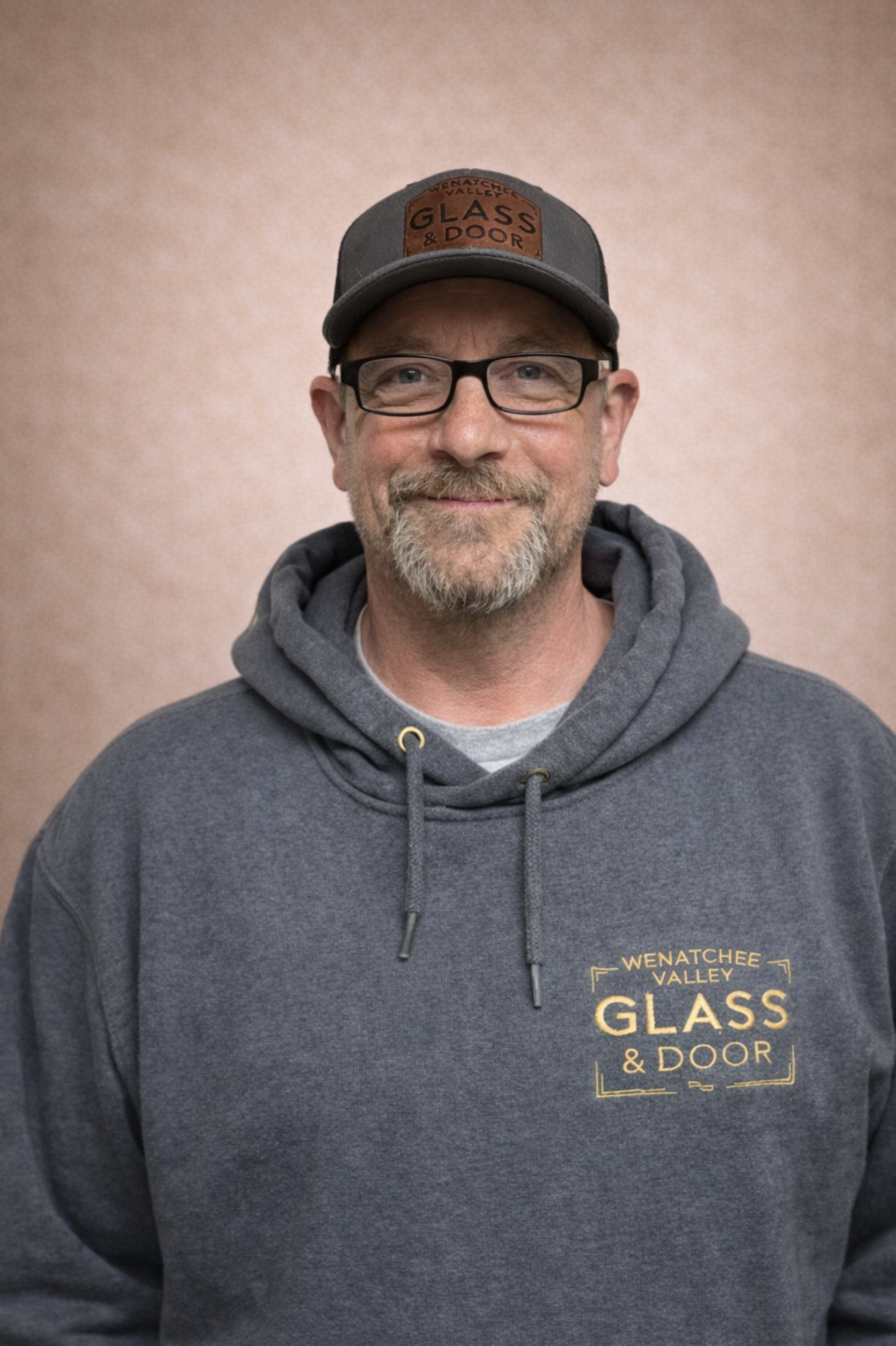 A smiling man with glasses and a beard wearing a gray hoodie and a gray cap with a leather patch that reads 'Glass & Door.' He stands against a beige background and the hoodie has a logo that reads 'Wenatchee Valley Glass & Door.'