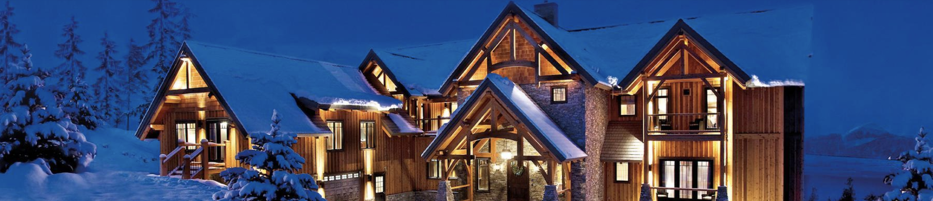 A large house with multiple gabled roofs illuminated at night, surrounded by snow-covered trees and landscape.