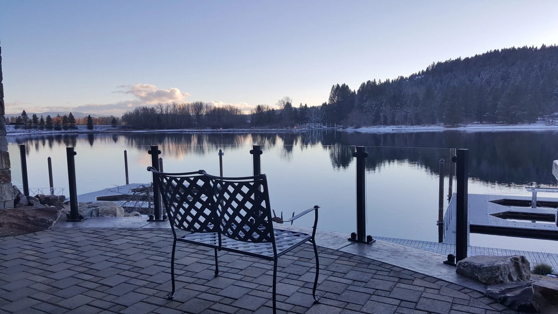 View of a calm lake surrounded by snow-covered trees with a stone and metal bench in the foreground on a brick patio, under a clear sky.