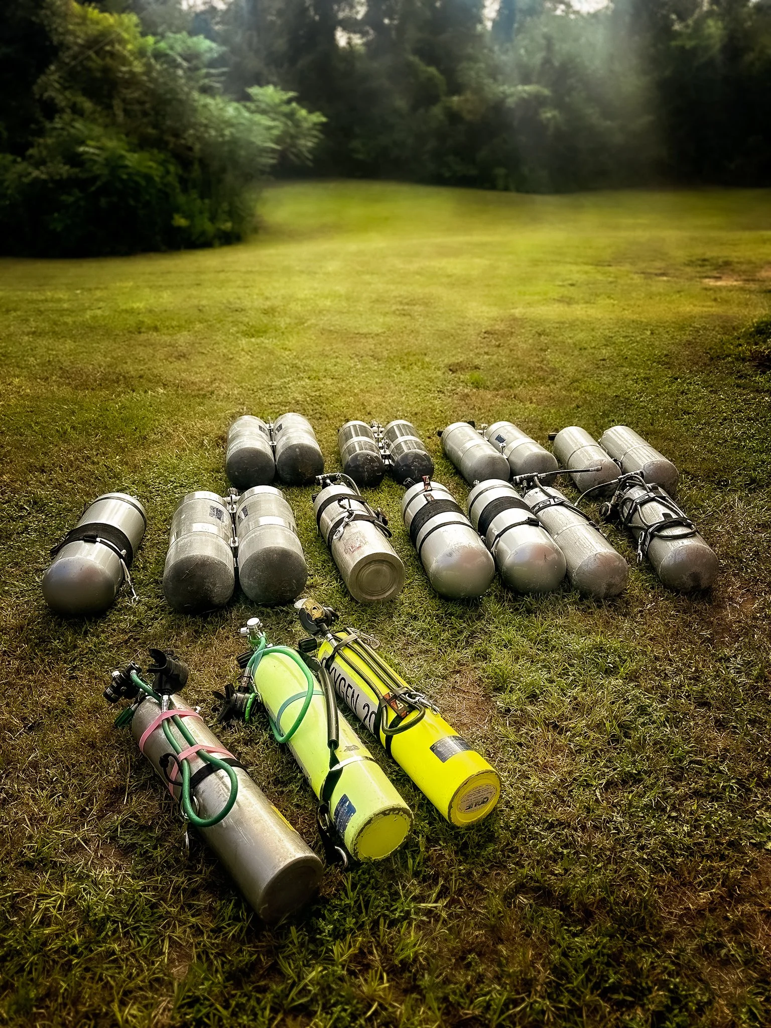 Multiple scuba tanks including sidemount, stage, and deco bottles with 100% oxygen lying on a grassy outdoor area with trees and a pathway in the background.