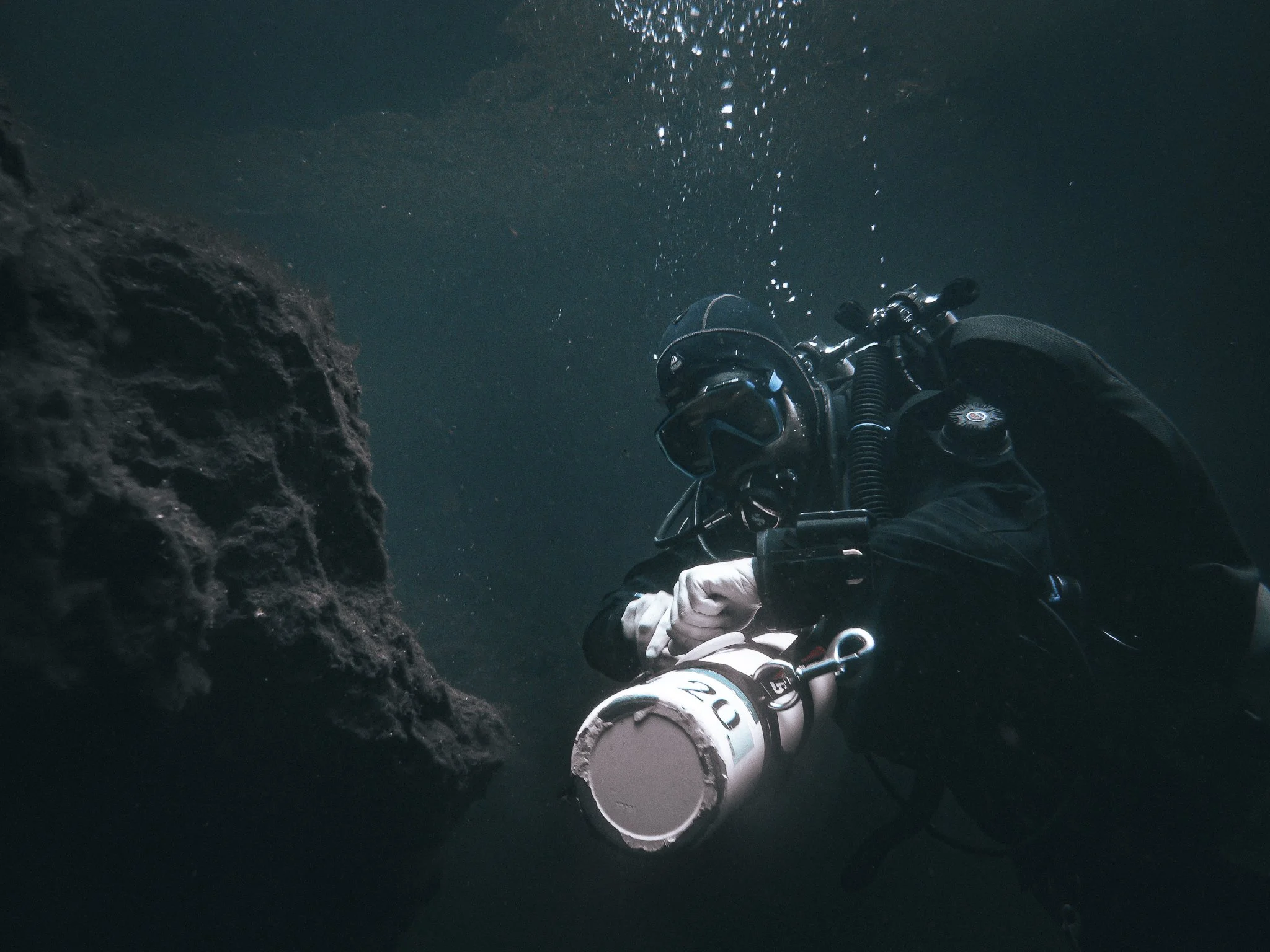 Underwater scene showing a diver wearing a helmet and suit, holding a large white tube or pipe, with dark water and a rocky underwater surface nearby.