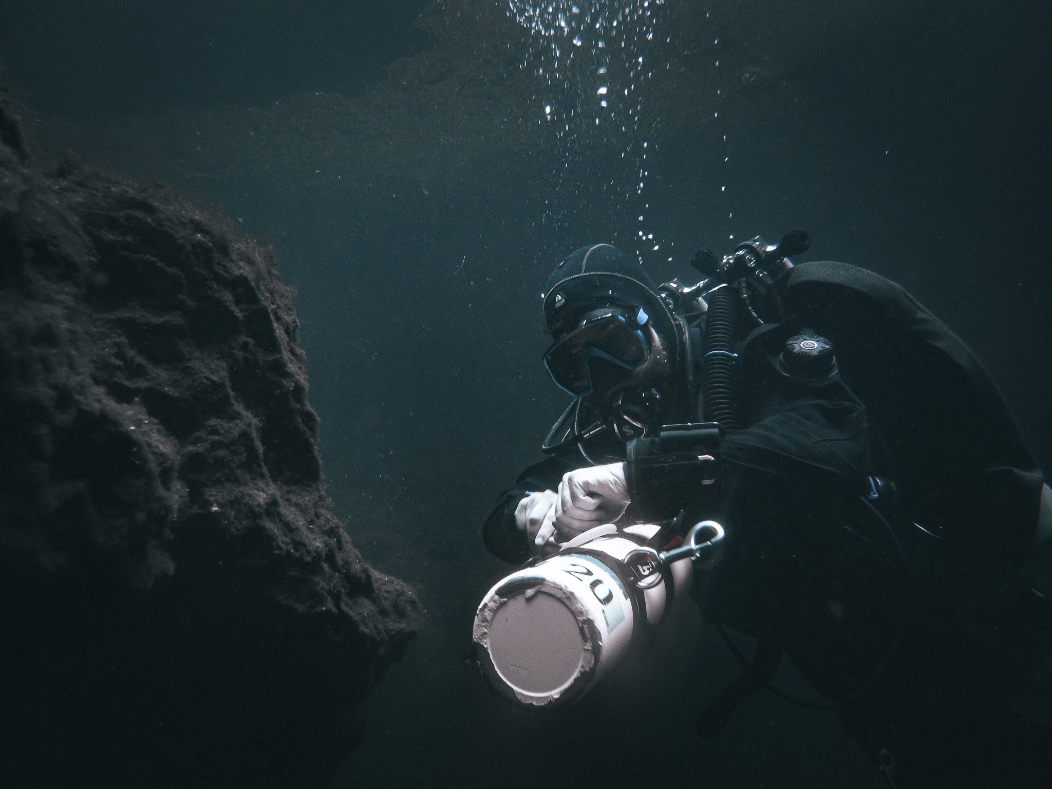 A diver underwater near a rocky surface, with water bubbles rising above.