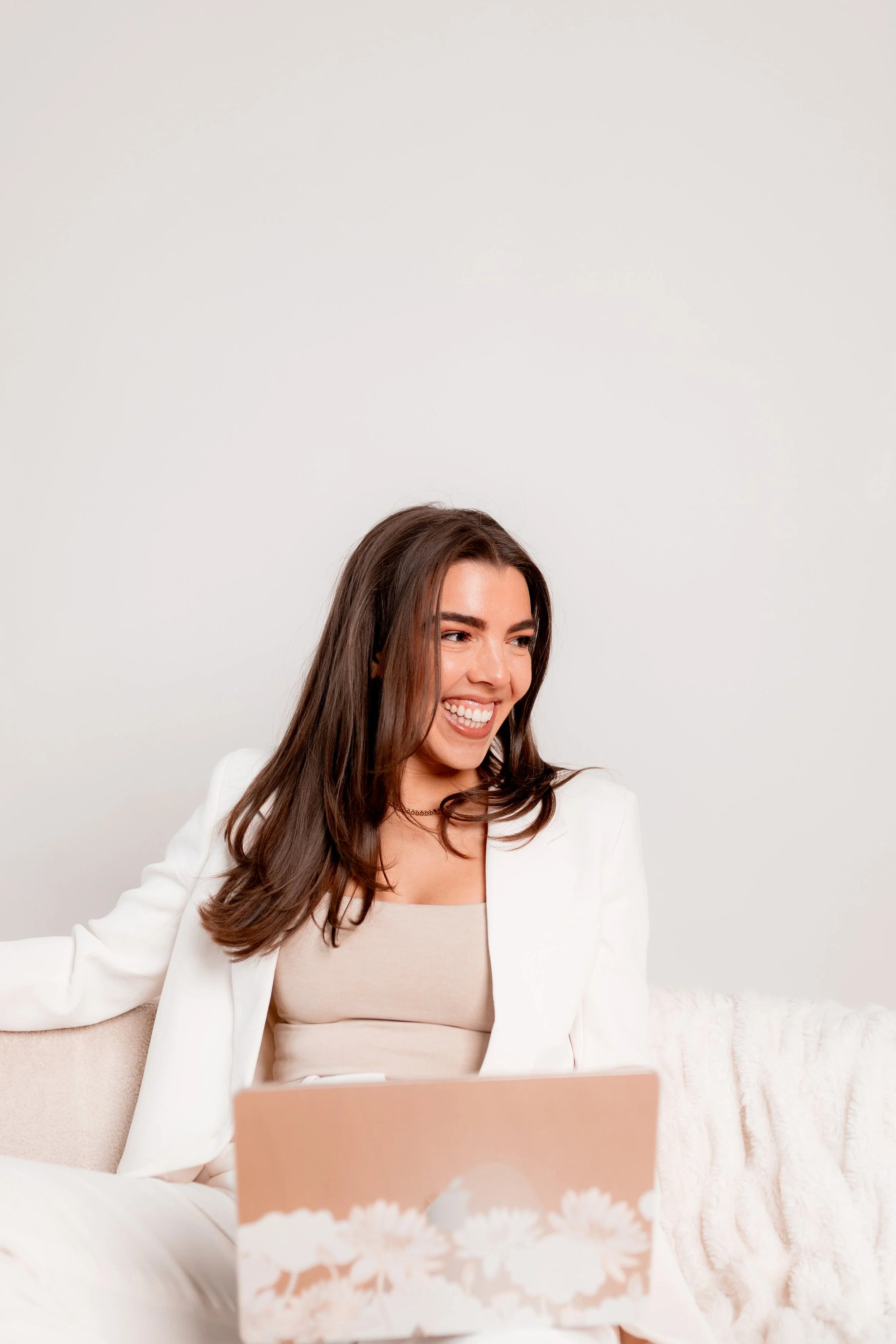 Happy woman sitting on a beige couch behind a floral gift bag, looking at a laptop.
