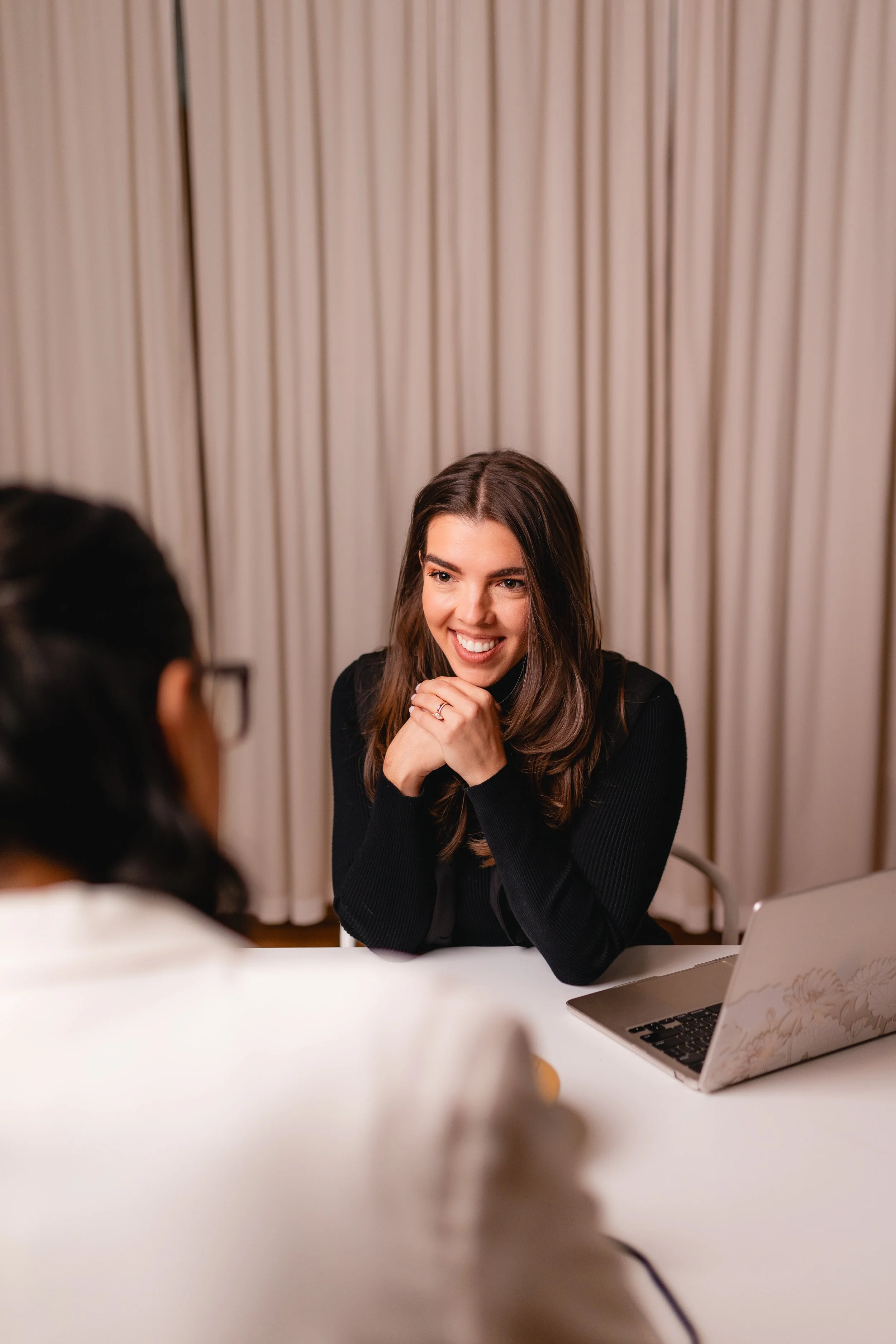 Smiling woman sitting at a table with a laptop, engaged in conversation or interview, in front of beige curtains.