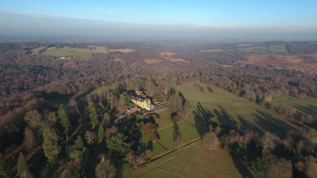 Aerial view of a pippingford estate surrounded by trees and open fields, with a castle-like building near the center, under a clear blue sky.