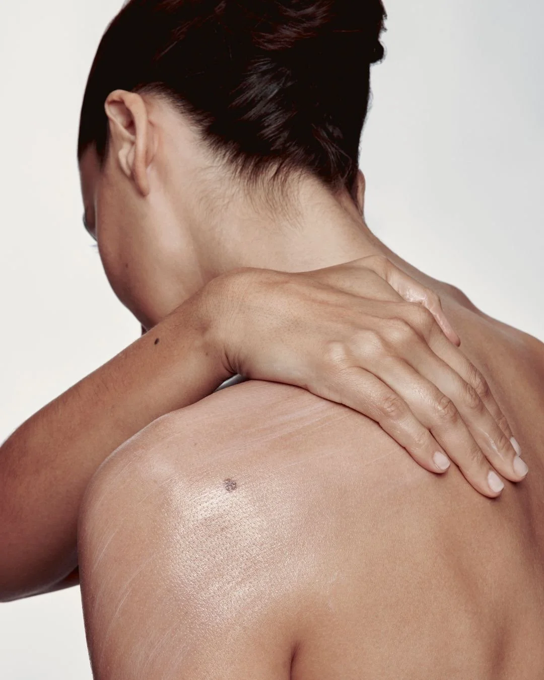 Close-up of a person with short dark hair, holding their shoulder with one hand, showing their back and neck, with a small mole on the shoulder, against a plain light background.