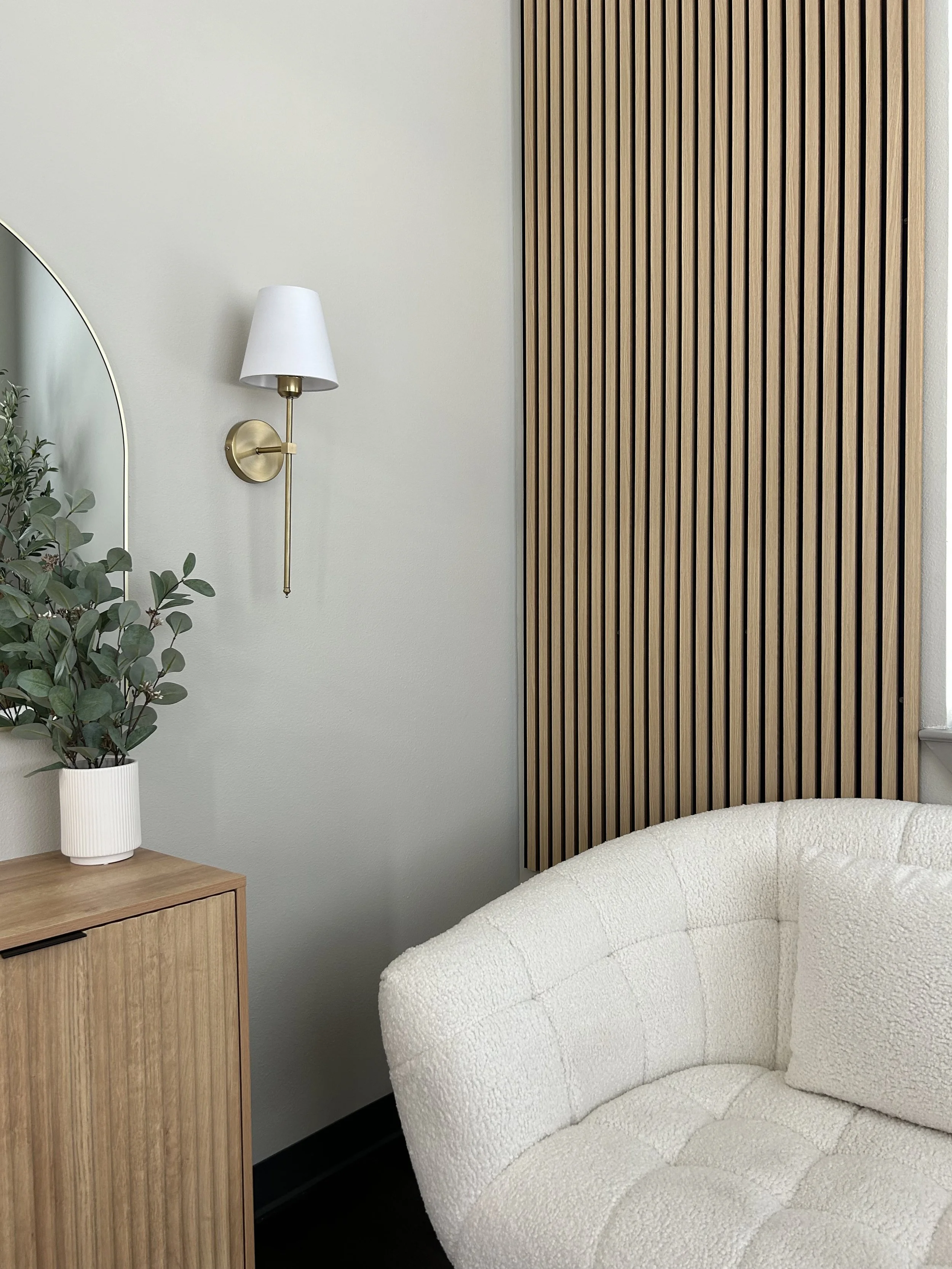 A close-up of a cozy corner of a room featuring a white textured armchair, a wooden side table with a white potted plant, a wall-mounted lamp with a white shade and brass arm, and a decorative wooden slat wall panel.