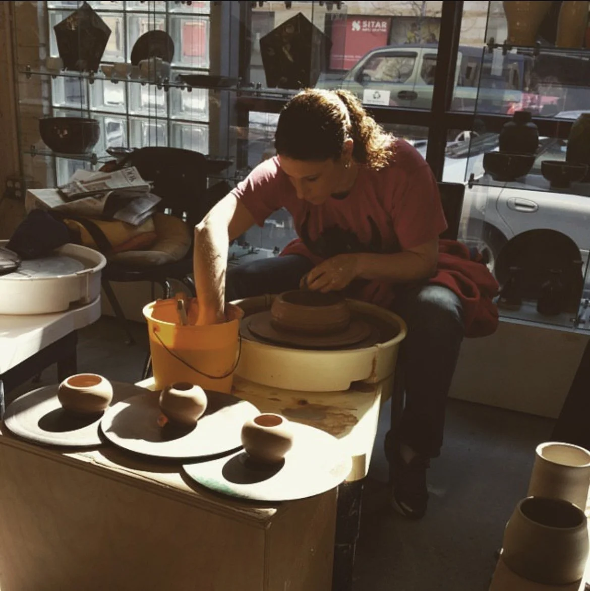 A woman working on pottery at a pottery wheel inside a pottery studio, with sunlight streaming through large windows. The studio has various pottery pieces and tools, with parked cars visible outside.