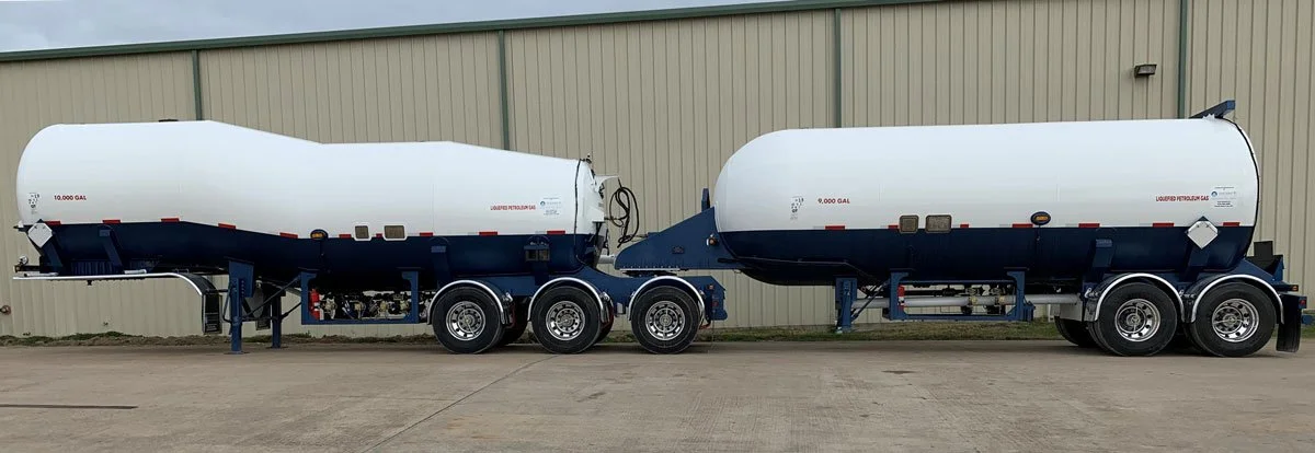 Two large tankers, used for transporting liquid fuel like gasoline, parked outside near a beige industrial building.