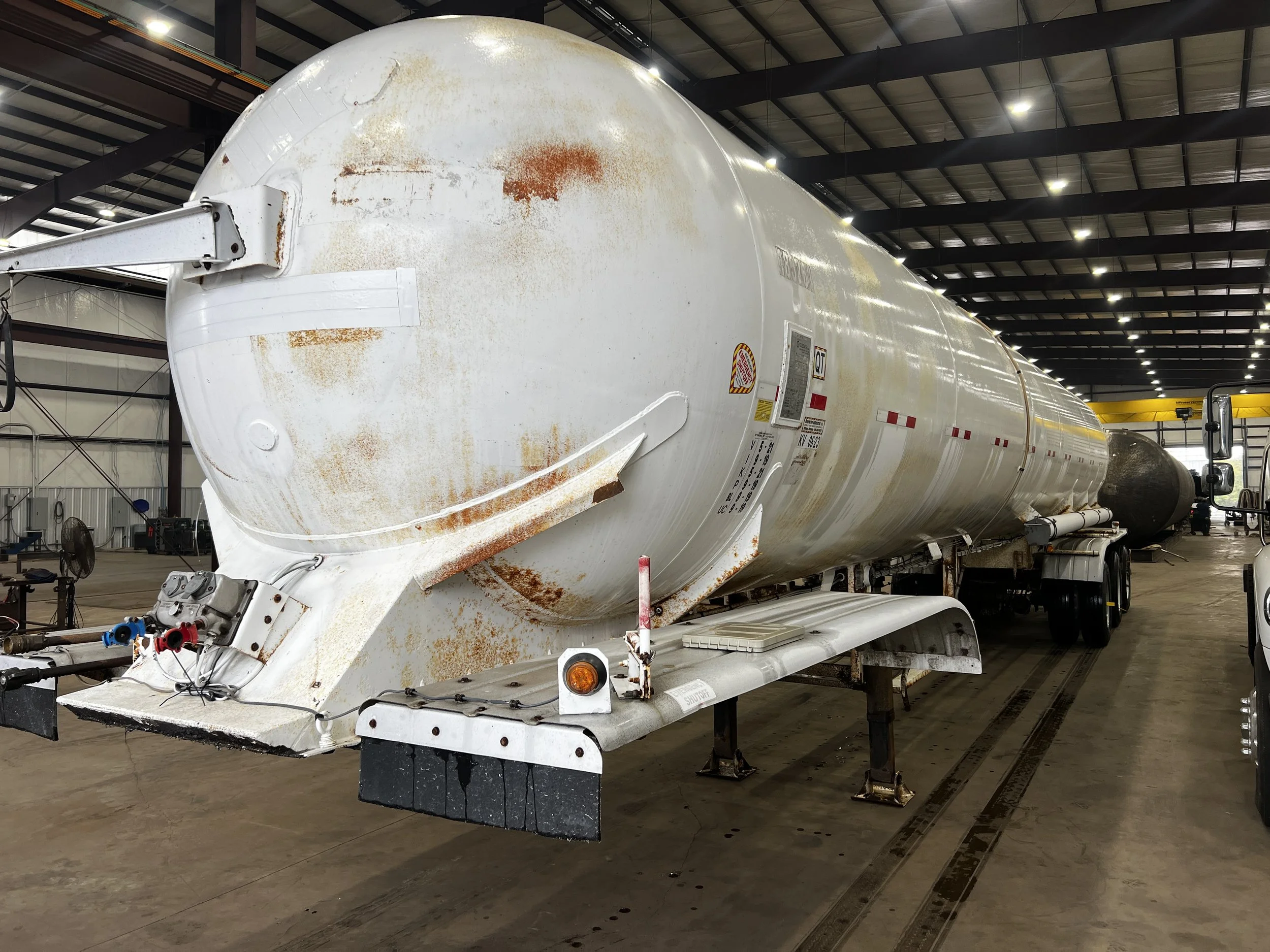 A large, white, cylindrical industrial storage tank with rust spots, sitting inside a spacious warehouse with a high ceiling and metal beams.