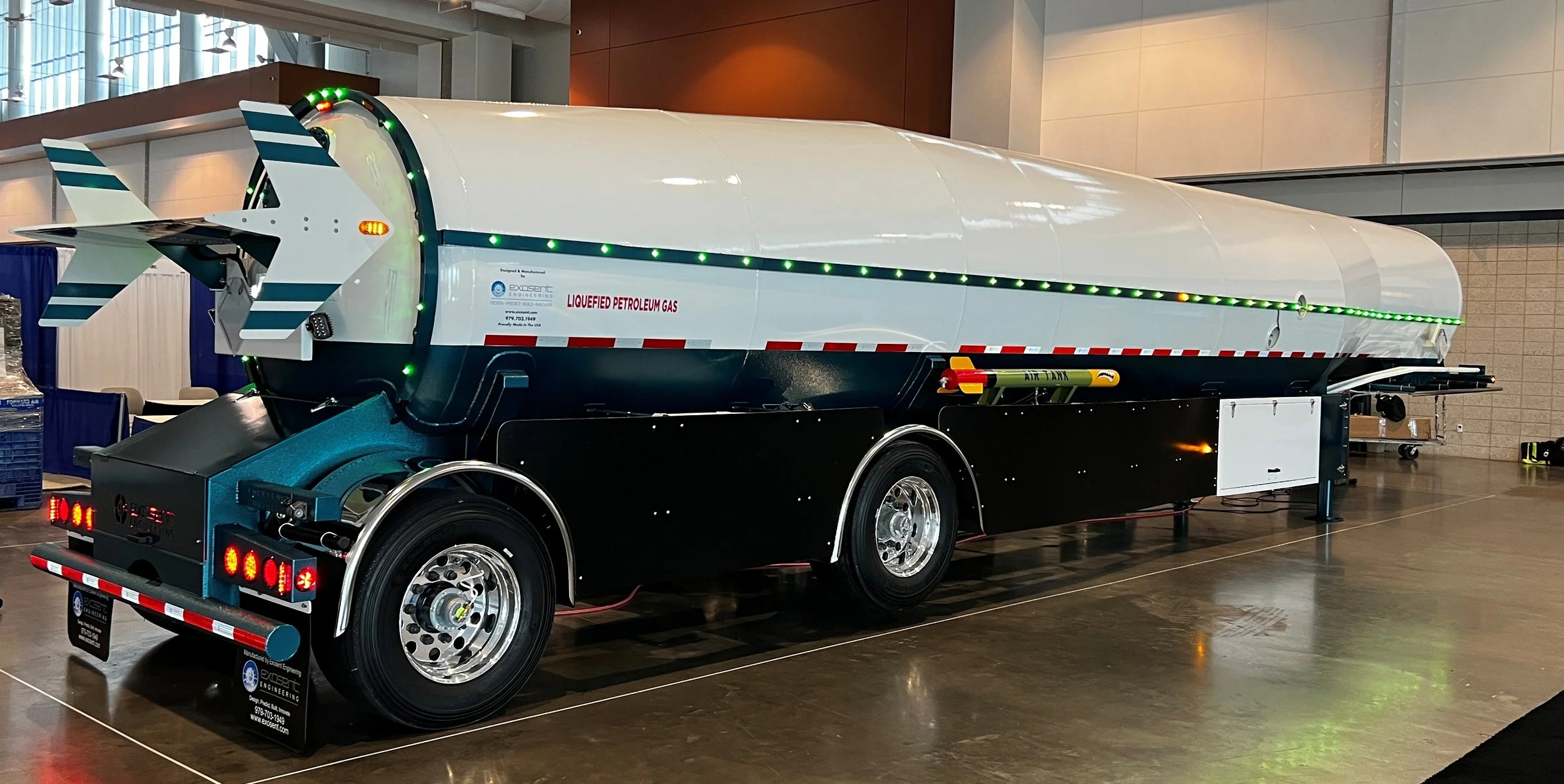 A large industrial liquefied petroleum gas (LPG) tank mounted on a black trailer with bright green and red LED lights, displayed indoors at an exhibition or conference.