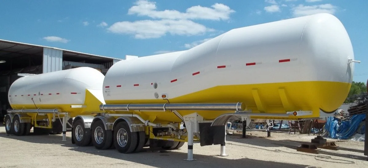 Two large white and yellow liquid fuel tankers parked outdoors on a gravel surface, with a warehouse and blue sky with clouds in the background.