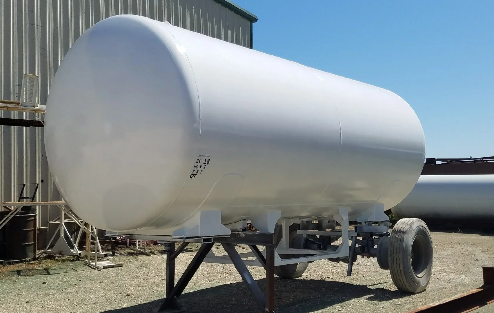 Large white cylindrical tank mounted on a trailer with a single axle and tire, outdoors near a metal building under a clear blue sky.