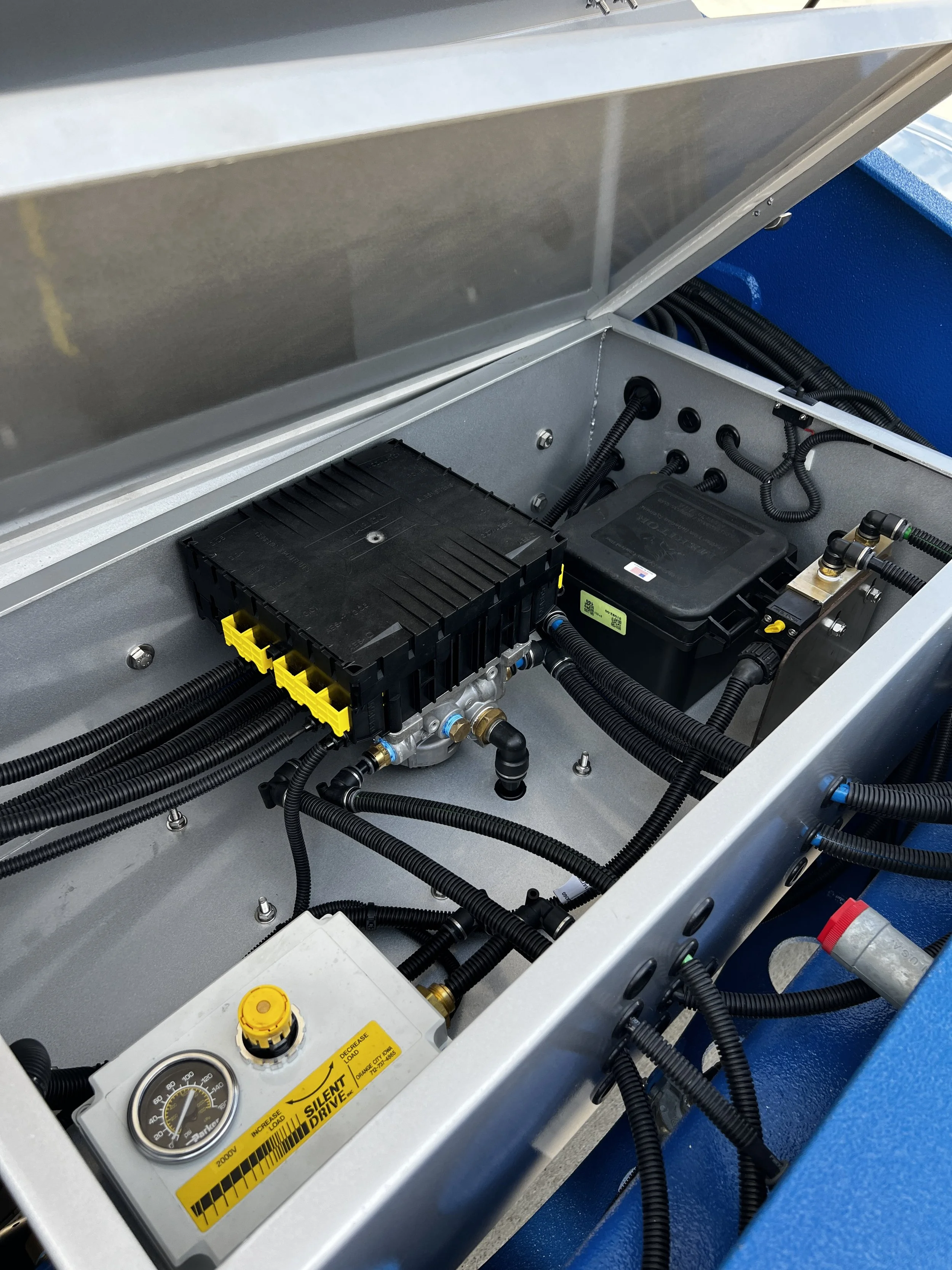 Interior view of a machine's electrical and hydraulic components, including black wiring, a gauge, and electronic modules inside a metal enclosure.