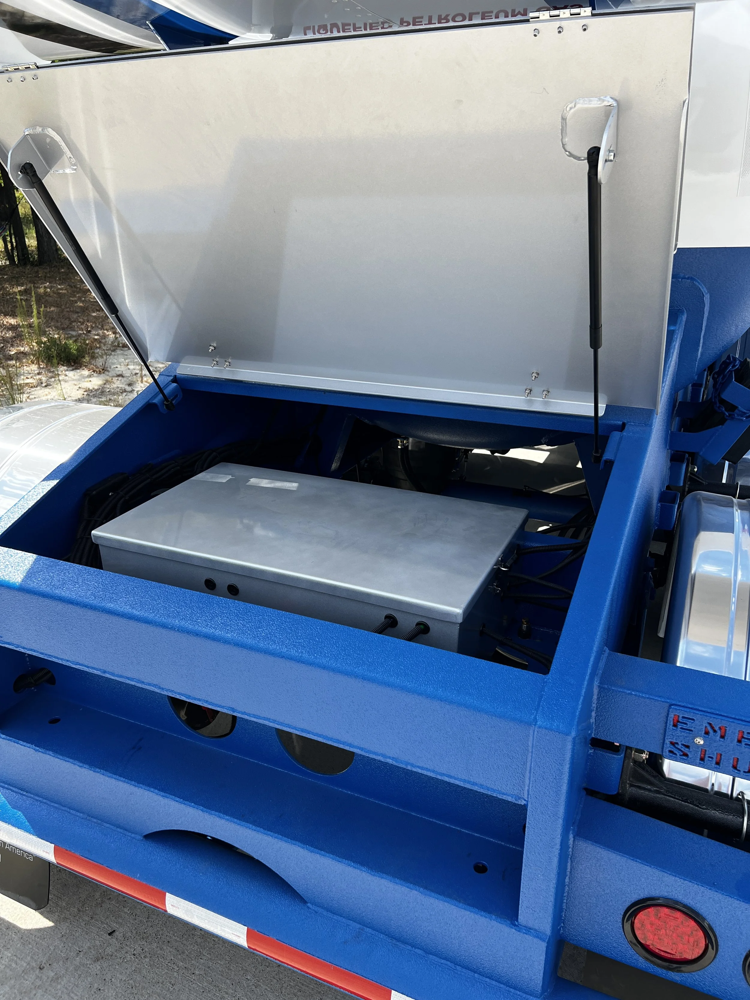 Open electric vehicle charging station compartment with wiring and a metallic box inside, blue exterior, and hydraulic supports holding the lid up.