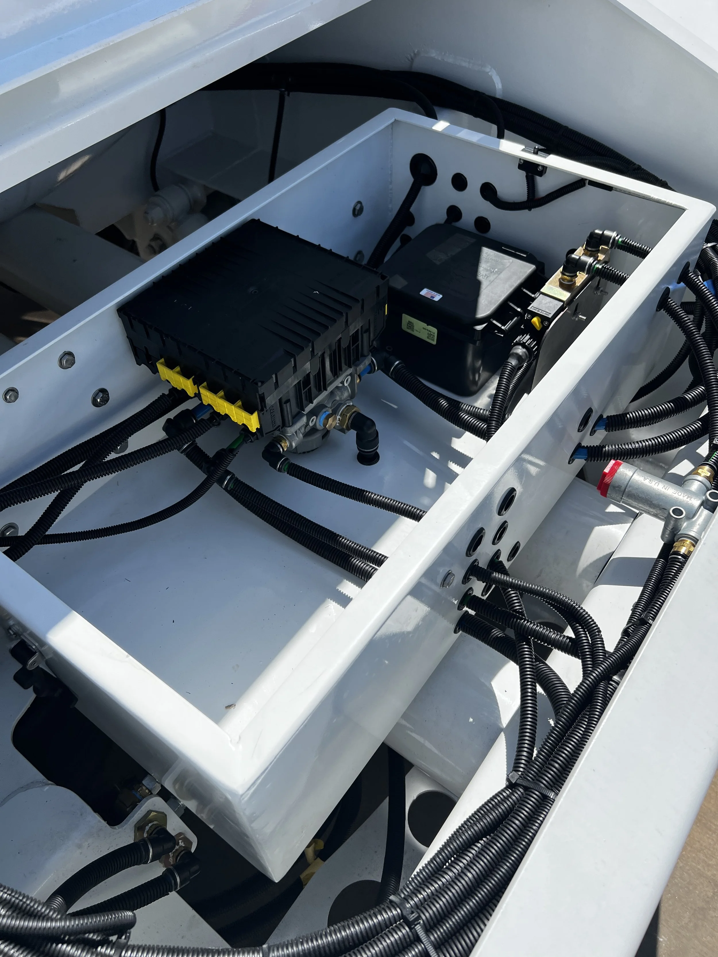 Electrical and hydraulic components with black wiring and tubing installed inside a white metal enclosure on a machine or vehicle.