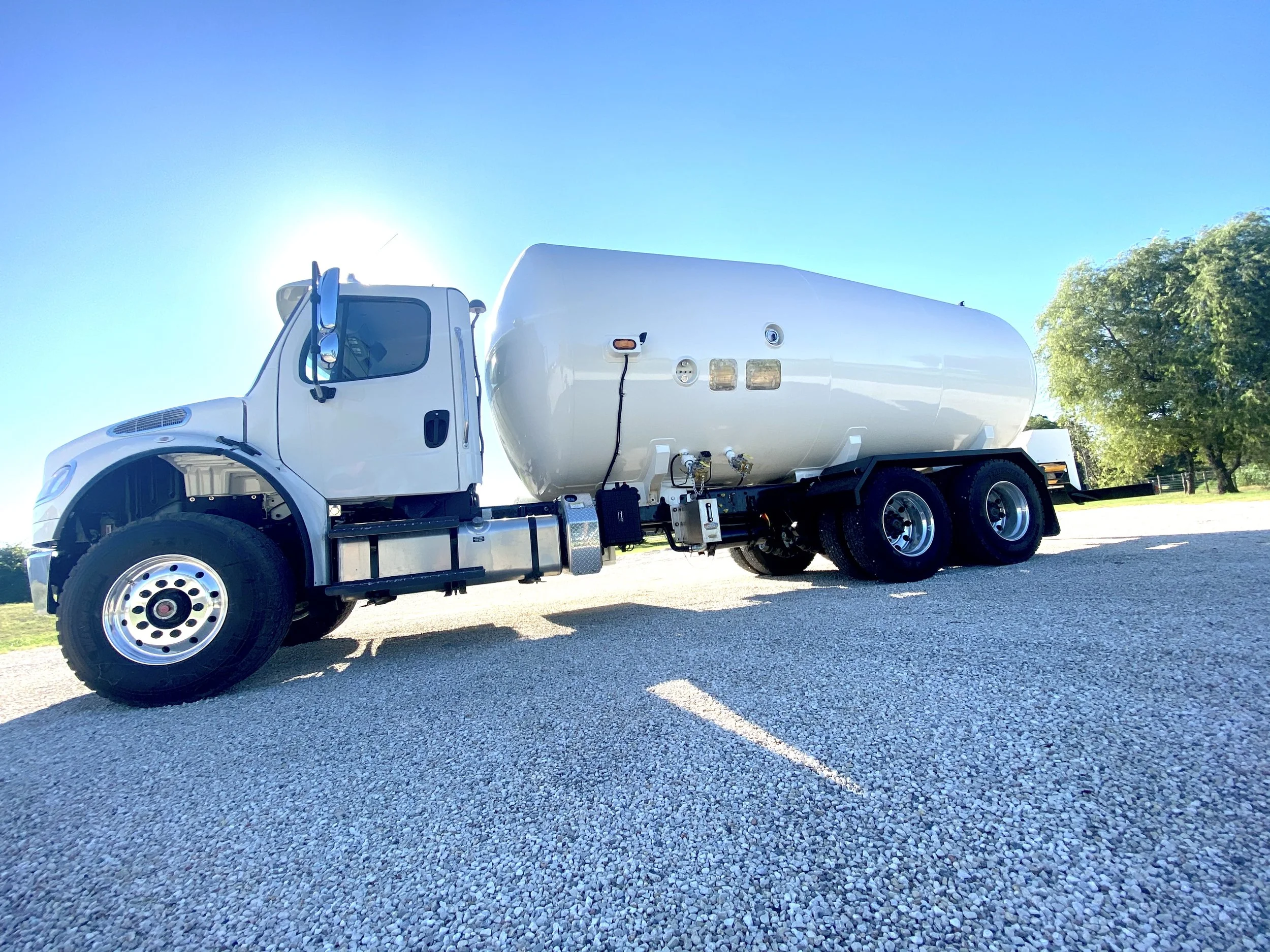White tank truck parked on gravel under clear blue sky with green trees in background.