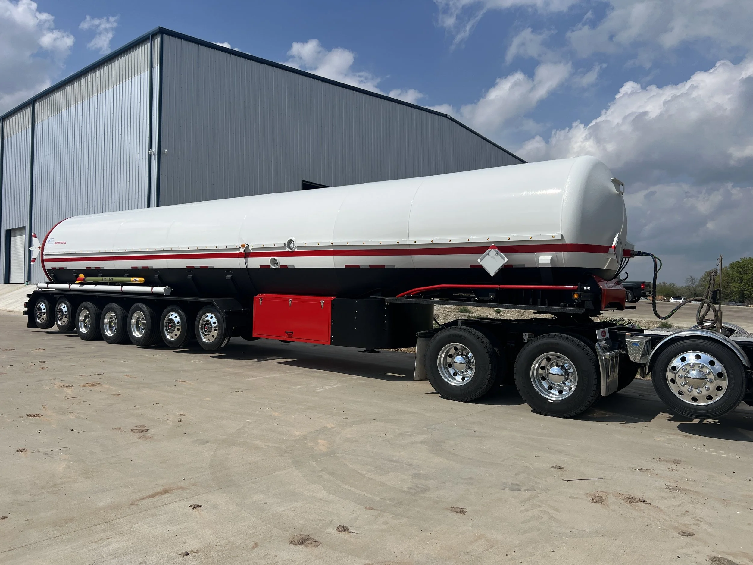 A large white tanker trailer with a red stripe parked on a concrete lot near a blue industrial building under a partly cloudy sky.