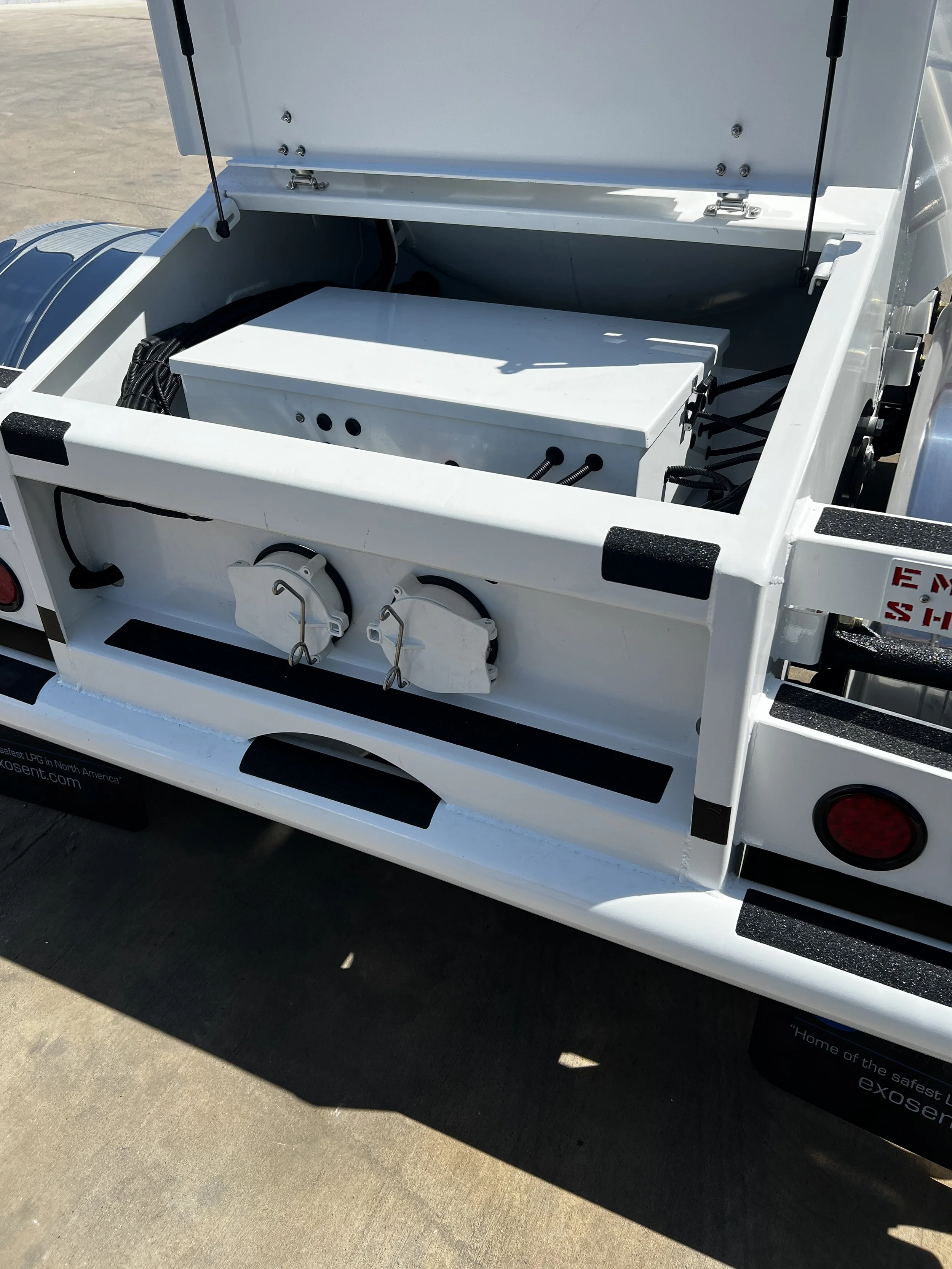 Open storage compartment of a white utility vehicle, revealing electrical components and wiring inside.