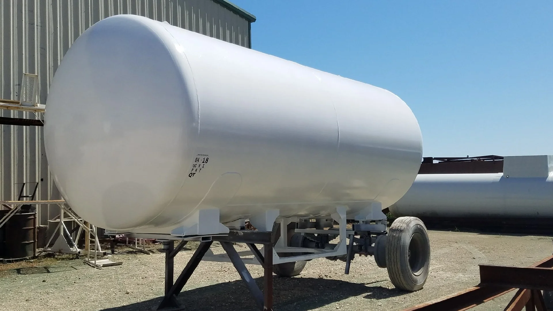 Large white cylindrical storage tank mounted on a trailer with large wheels, outdoors on a gravel surface under a clear blue sky.