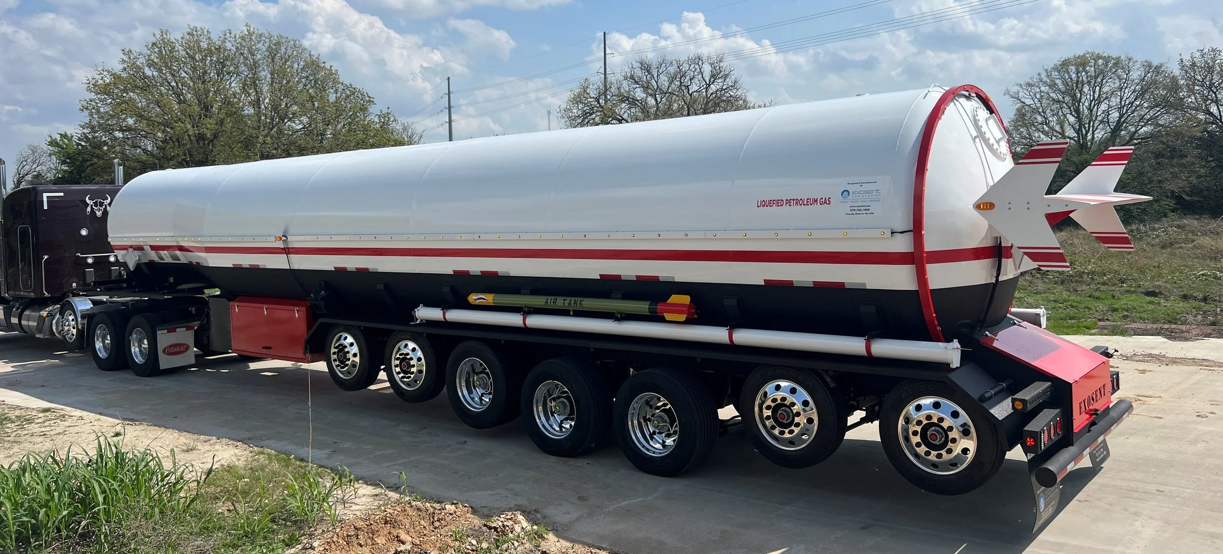 A fuel tanker with a missile-shaped design on the trailer, labeled for liquefied petroleum gas, parked on a concrete surface outdoors with trees and a cloudy sky in the background.